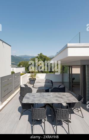 Balcone esterno della villa modena con un piccolo soggiorno composto da divano, due poltrone e un tavolo con sedie. Sullo sfondo colline svizzere Foto Stock