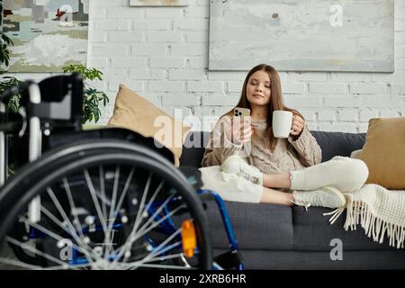 Una giovane donna su una sedia a rotelle si rilassa sul divano, gustando una tazza di caffè e navigando sul telefono. Foto Stock