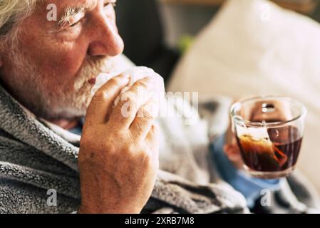 Un anziano con influenza fredda sintomi influenzali bere medicina o tè alle erbe naturale a casa per guarire. Concetto di virus e anziani maturi con problemi di salute. Persone e malattie invernali Foto Stock