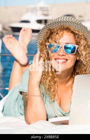 Giovane bella donna adulta sorridere e utilizzare computer portatile esterno posare sul ponte di uno yacht - viaggio e vacanza estiva stile di vita e moderno lavoro gratuito - lavoratori intelligenti Foto Stock