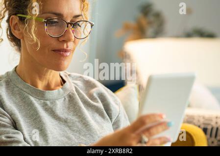 Primo piano di donna moderna donna adulta che legge un libro da un dispositivo di lettura elettronico tablet. Signora che indossa occhiali da vista e sorride godendosi un po' di relax a casa seduto sul divano Foto Stock