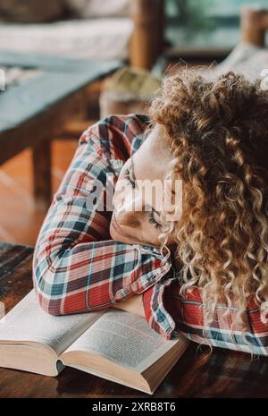 Donna carina che dorme su un libro aperto da sola a casa. Donne stanche che dormono sul tavolo. Concetto di stile di vita degli studenti. Una bella bionda lunga e riccia che riposa nel soggiorno. Abitacolo accogliente Foto Stock