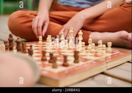 Uno scatto ravvicinato di due persone che partecipano a una partita a scacchi su una tavola di legno all'aperto. L'immagine si concentra sui pezzi e sulle mani degli scacchi, evidenziando la S Foto Stock