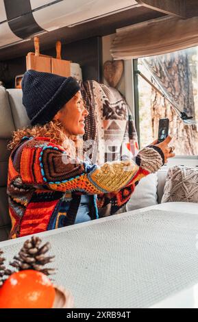 Donna che indossa un cappello e un maglione da pacwork seduto in camper e scatta foto con il cellulare che guarda fuori dalla finestra. Concetto di relax e svago Foto Stock