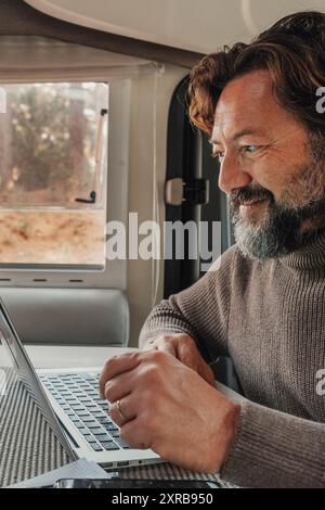 Un uomo lavora su un computer portatile all'interno di un camper seduto al tavolo della scrivania. Stile di vita moderno del lavoro di connessione online. Persone e nomade digitale van vita lifestyle. Natura all'aperto fuori dalla finestra. Libertà Foto Stock