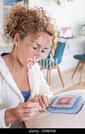 Donna caucasica adulta a casa che svolge attività di hobby all'uncinetto a casa decorando un cuscino bianco shabby chic. Interno del soggiorno sullo sfondo. Donne che creano decorazioni di lana Foto Stock