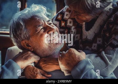 Ritratto di una coppia di anziani innamorati a casa alla luce buia della sera. Il vecchio sorride alla sua donna matura, moglie che abbraccia e si gode la relazione. Home background invernale. La vita degli anziani Foto Stock