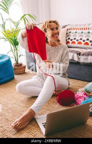 Una donna felice a casa mostra i risultati del lavoro a maglia. Laptop sul tappeto. Hobby online Internet attività ricreative persone. Donna adulta che si diverte al chiuso. Lavorare a maglia allegro stile di vita femminile Foto Stock