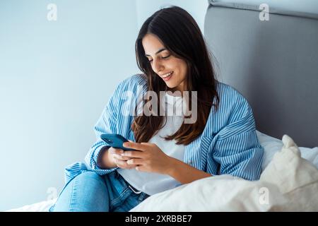 Giovane donna seduta sul letto, vestita con abiti casual, tenendo il telefono e leggendo nuovi messaggi, sorridendo pacificamente. Concetto tecnologico. Foto Stock