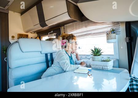 Una donna si rilassa all'interno di un camper camper. la signora ama rilassarsi sedendosi sul divano guardando fuori. Concetto di libertà di viaggio. Stile di vita femminile viaggiatore nel weekend di primavera. Indipendenza Foto Stock