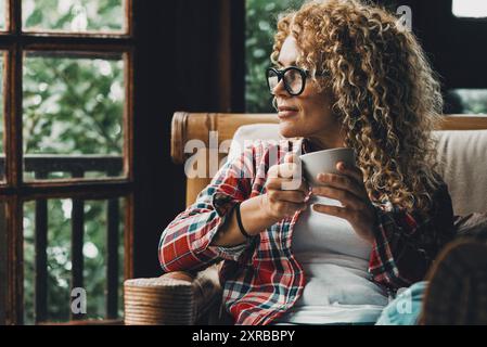 Una donna attraente di mezza età che si rilassa a casa seduto sulla sedia e guarda all'esterno, godendo del tempo pomeridiano e delle attività ricreative al coperto da sola. Donne indipendenti al chiuso. rilassatevi Foto Stock