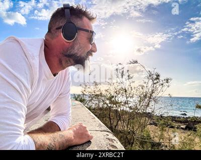 Un uomo maturo che indossa le cuffie che ascolta musica sulla costa godendosi l'oceano di sottofondo. ahd per turisti attività ricreative all'aperto persone mature. Rilassatevi e stile di vita maschile sereno Foto Stock