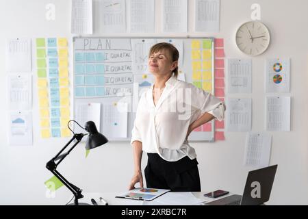 Donna anziana che soffre di mal di schiena dopo il lavoro per molto tempo che utilizza un computer portatile sul posto di lavoro. Schiena bassa, mal di vita, lumbago e reumatismi. Sindrome da ufficio Foto Stock