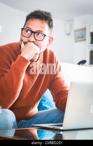 Serio uomo caucasico maturo con barba che pensa mentre si siede sul divano nel suo moderno salotto di lavoro. Concetto di persone aziendali che utilizzano dispositivi mobili. Sfondo sfocato. Uomini premurosi Foto Stock