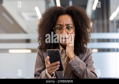 Giovane donna d'affari afro-americana che guarda scioccata davanti allo smartphone in un moderno ambiente d'ufficio. La donna appare sorpresa da notizie o messaggi inaspettati al telefono mentre lavora Foto Stock
