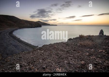 Vista dell'alba sull'Atlantico. Una costa e una baia con una spiaggia rocciosa su un'isola vulcanica. Pietre laviche fredde in riva al mare presso la spiaggia Playa Laja del Corral, Fuerteventura, Isole Canarie, Spagna Foto Stock