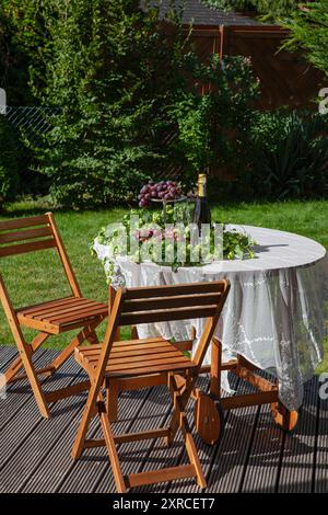 Un accordo con 2 bicchieri da champagne e una bottiglia di champagne accanto all'uva rossa e al luppolo di birra come decorazione, due sedie di legno si trovano accanto a un tavolo con tovaglia bianca su una terrazza in un giardino, per preparare una festa in giardino, festività e celebrazioni nel verde e all'aperto Foto Stock