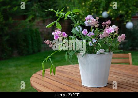 Un cachepot bianco con fiori freschi come i sacchetti rosa, le vecce bianche e rosa e le foglie verdi si erge come una disposizione floreale su un tavolo di legno in primo piano; un albero di mele con mele verdi chiare si staglia sfocato sullo sfondo del giardino Foto Stock