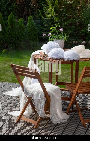 Abiti e decorazioni bianchi festivi adagiati su un tavolo di legno su una terrazza nel giardino, per preparare un matrimonio, sfondo verde sfocato nel giardino Foto Stock