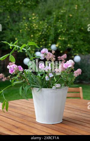 Un cachepot bianco con fiori freschi da giardino come i sacchetti rosa, vetche bianche e rosa e foglie verdi si erge come una disposizione floreale su un tavolo di legno nel giardino; un albero di mele con mele verdi chiare si staglia sfocato sullo sfondo Foto Stock