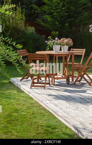 Mobili da giardino su una terrazza in legno nel verde giardino, fiori fioriti in due vasi bianchi come tovaglia nell'oasi del giardino, foglie di salice come ombra, vacanze a casa in estate, Foto Stock