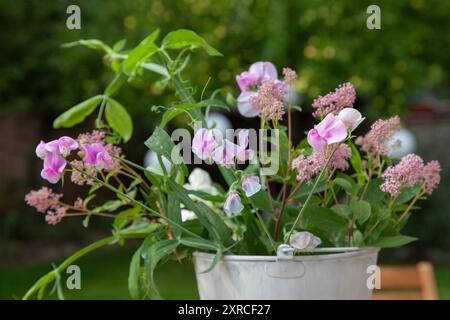 Un cachepot bianco con fiori freschi come i sacchetti rosa, le vecce bianche e rosa e le foglie verdi si erge come una disposizione floreale su un tavolo di legno in primo piano; un albero di mele con mele verdi chiare si staglia sfocato sullo sfondo del giardino Foto Stock