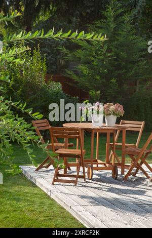 Mobili da giardino su una terrazza in legno nel verde giardino, fiori fioriti in due vasi bianchi come tovaglia nell'oasi del giardino, foglie di salice come ombra, vacanze a casa in estate, Foto Stock