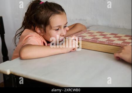 Una giovane ragazza appare profonda nel pensiero e concentrata mentre gioca a un gioco da tavolo al chiuso. Appoggia la testa sul tavolo, pensando alla sua prossima mossa. Foto Stock