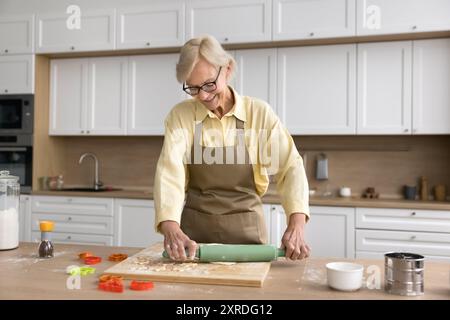 Una donna anziana bionda felice che prepara biscotti a casa, impasto arrotolato Foto Stock