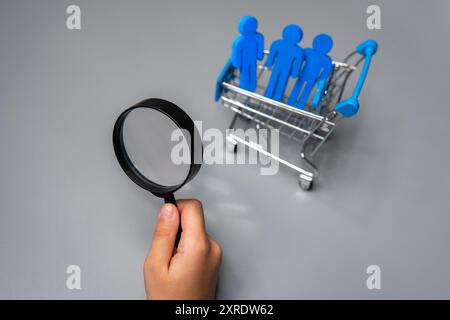 Una mano che regge una lente d'ingrandimento e un carrello della spesa in miniatura pieno di statuette blu. Ricerca dei clienti, concetto di segmentazione del mercato. Foto Stock