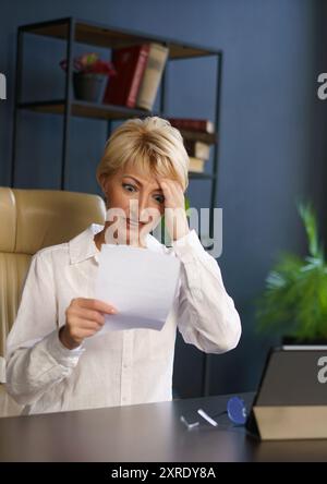 Una donna appare ansiosa mentre legge un documento, seduto alla sua scrivania con un tablet elettronico nelle vicinanze in un ambiente di ufficio contemporaneo. Foto Stock