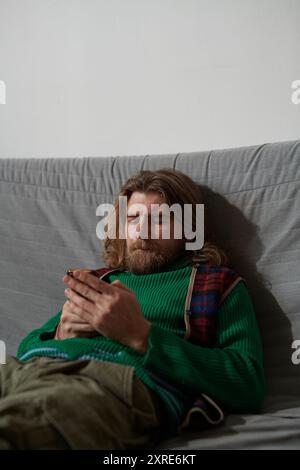 Uomo barbuto che indossa un maglione verde e una sciarpa a quadri seduto sul divano e che digita al telefono, mostrando un'espressione mirata senza guardare la fotocamera Foto Stock
