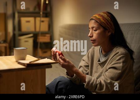 Donna seduta in una stanza accogliente che tiene in mano uno smartphone rosso con un'espressione premurosa e che guarda lo schermo Foto Stock