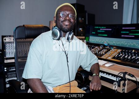 Uomo seduto in studio di registrazione con le cuffie intorno al collo, sorridente. Lo sfondo presenta varie apparecchiature audio, console di mixaggio e schermi di computer Foto Stock