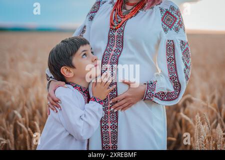 Mamma Ucraina incinta con figlio in campo di grano, sulla natura. Felice sorridente fratello ragazzo toccando mamme bump pancia. Foto Stock