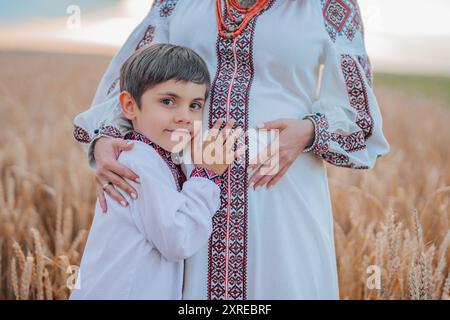 Mamma Ucraina incinta con figlio in campo di grano, sulla natura. Felice sorridente fratello ragazzo toccando mamme bump pancia. Foto Stock