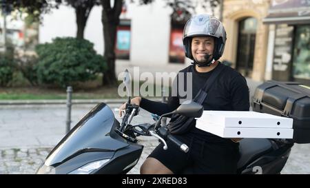 Un uomo che indossa un casco sta guidando una motocicletta con una scatola per pizza sul retro. Sta sorridendo e si sta godendo il suo passaggio Foto Stock