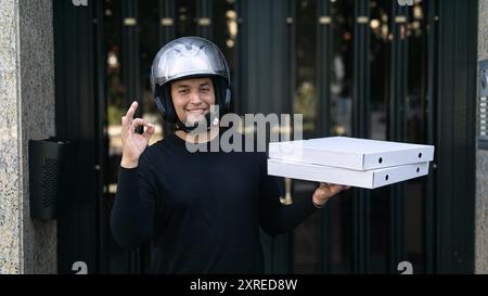 Un uomo con un casco ha due scatole per pizza. Sta sorridendo e alzando la mano nel segno giusto. Foto Stock