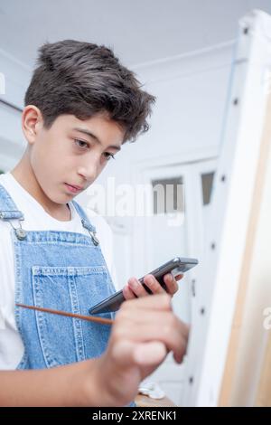 Ragazzo adolescente che dipinge su una tela con un pennello mentre consulta il suo telefono cellulare. Foto Stock