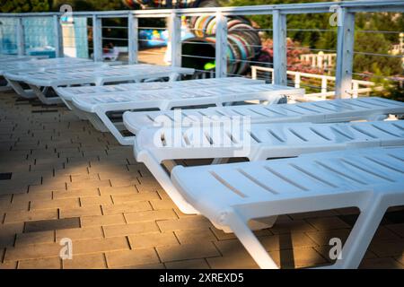 Lettini a bordo piscina - lettini in plastica bianchi allineati su un ponte con una piscina sullo sfondo. Foto Stock