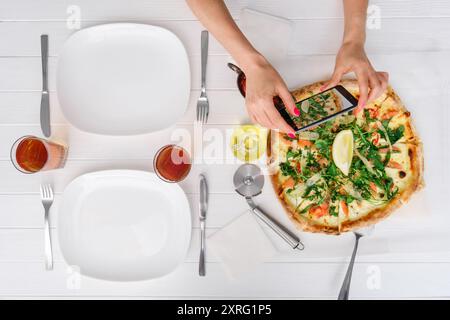 La donna scatta una foto di una pizza fresca prima di mangiare su un tavolo di legno bianco Foto Stock