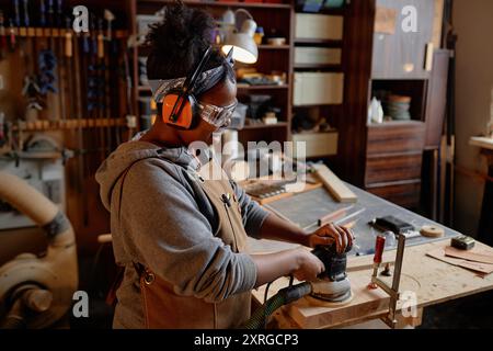 Giovane donna nera in officina levigatura industriale del legno con attrezzatura protettiva circondata da utensili e materiali per la lavorazione del legno Foto Stock