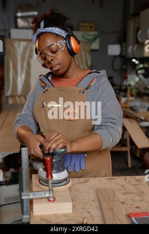 Donna afroamericana che leviga pezzi di legno in officina indossando indumenti protettivi, tra cui occhiali e cuffie che mostrano concentrazione e dedizione Foto Stock