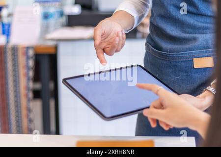 Una giovane donna che possiede un bar e un barista utilizza un tablet per prendere ordini da una cliente femminile che si trova da sola all'interno del negozio. Self-relian Foto Stock