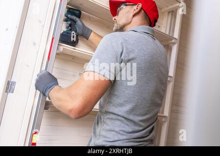 Un carpentiere esperto si concentra sull'installazione di scaffali in un'officina ben illuminata, utilizzando un utensile elettrico per la precisione. Foto Stock