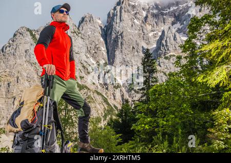 Un escursionista con una giacca rossa si erge con sicurezza su un affioramento roccioso, circondato da torreggianti montagne e alberi lussureggianti. Foto Stock