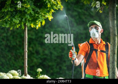 Un giardiniere in una camicia arancione e equipaggiamento protettivo spruzza pesticidi sulle piante, garantendo la salute del giardino. Foto Stock