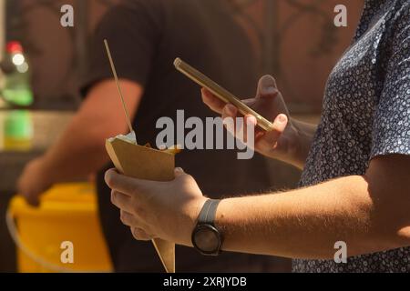 Metà di un uomo che tiene un cono con patatine fritte in una mano, smartphone nell'altra mano e scatta foto del suo cibo. Foto Stock