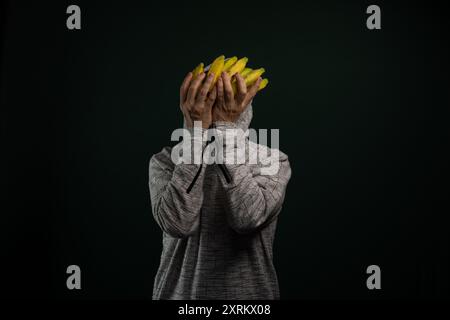 Uomo misterioso che indossa una felpa con cappuccio che tiene un mucchio di banane gialle davanti al viso. Isolato su sfondo scuro. Foto Stock