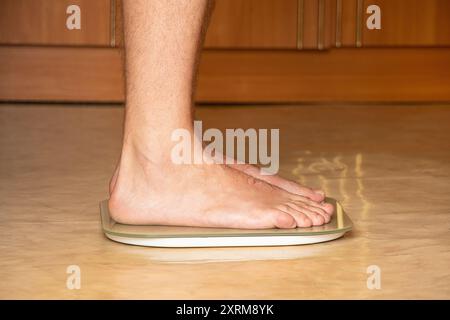 Un uomo sta in piedi su una scala sul pavimento di un appartamento, monitorando il suo peso, a dieta Foto Stock